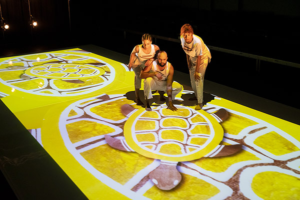 Three indigenous performers stand on an art digital projection