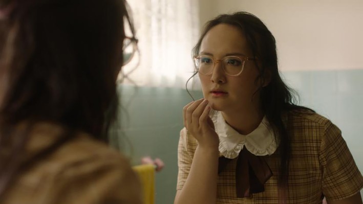 A woman with glasses in a ruffled blouse and plaid vest attentively looks at her reflection in a mirror, her expression thoughtful and introspective.