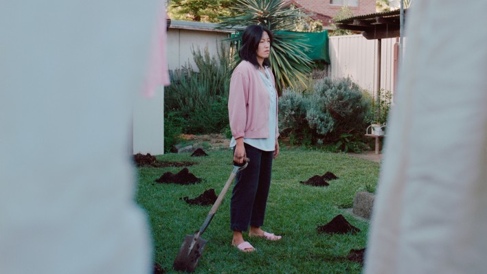 A woman stands in a garden holding a shovel, surrounded by patches of dug-up earth. She wears a casual outfit with a pink jacket and jeans, appearing contemplative as she gazes off to the side.