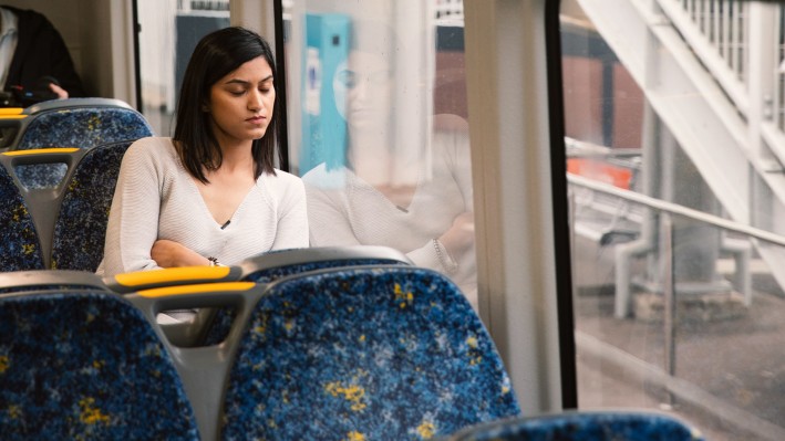 A contemplative woman sits alone on a train, gazing downward with a notebook on her lap, framed by the window reflecting her image and the passing outdoor scene.