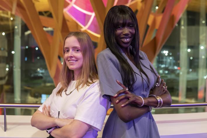 Two women stand back to back, facing the camera and smiling