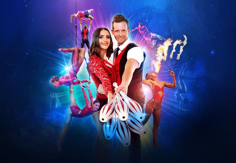 A woman in a red dress stands behind a man in a red vest, with Cirque Nouvelle performers (acrobats) in the background