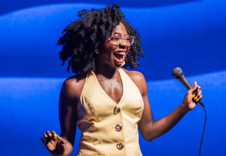 A woman in a yellow dress against a blue background, smiling as she holds a microphone - Anisa Nanduala