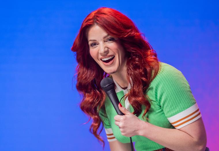 A woman with red hair, wearing a green dress, holds a microphone - Bron Lewis
