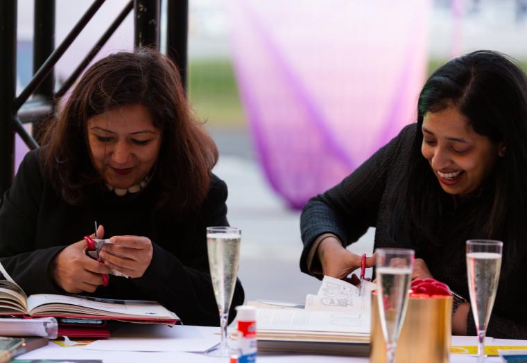 Two people sitting next to each other cutting out images, smiling and drinking wine