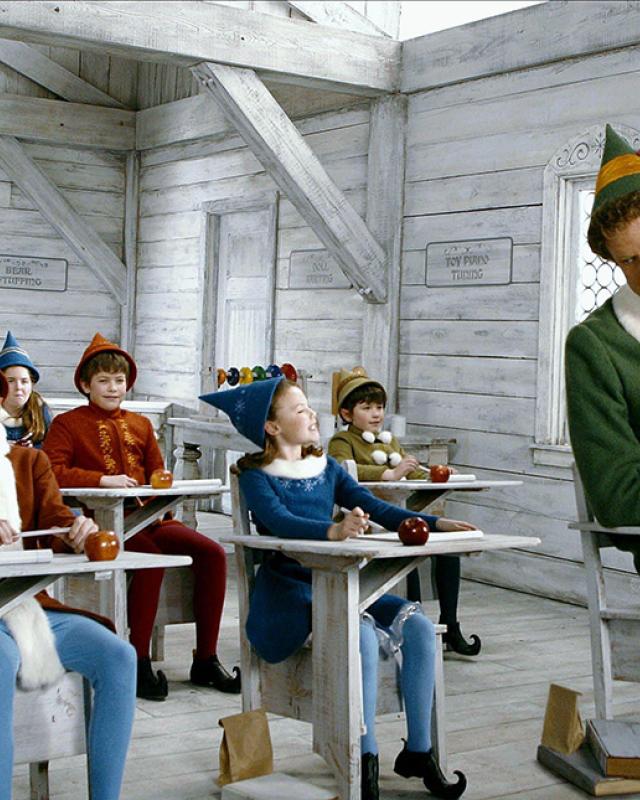 A large man dressed as an elf sits in a room full of smaller elves, from the movie Elf.
