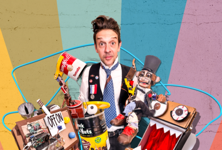 Mario the Maker Magician is a man in a vest surrounded by lots of rubbish including boxes and tin cans.