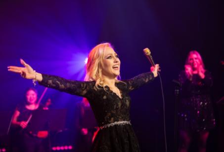 A blonder woman in a black dress holds her arms out on stage - To Be Loved - A Dedication to Adele