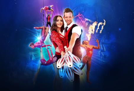 A woman in a red dress stands behind a man in a red vest, with Cirque Nouvelle performers (acrobats) in the background